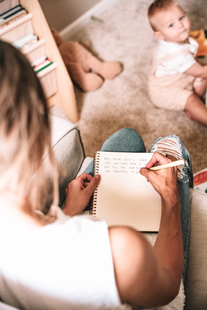 a woman sitting on a couch, writing in a notebook with a baby sitting on the floor beside her.